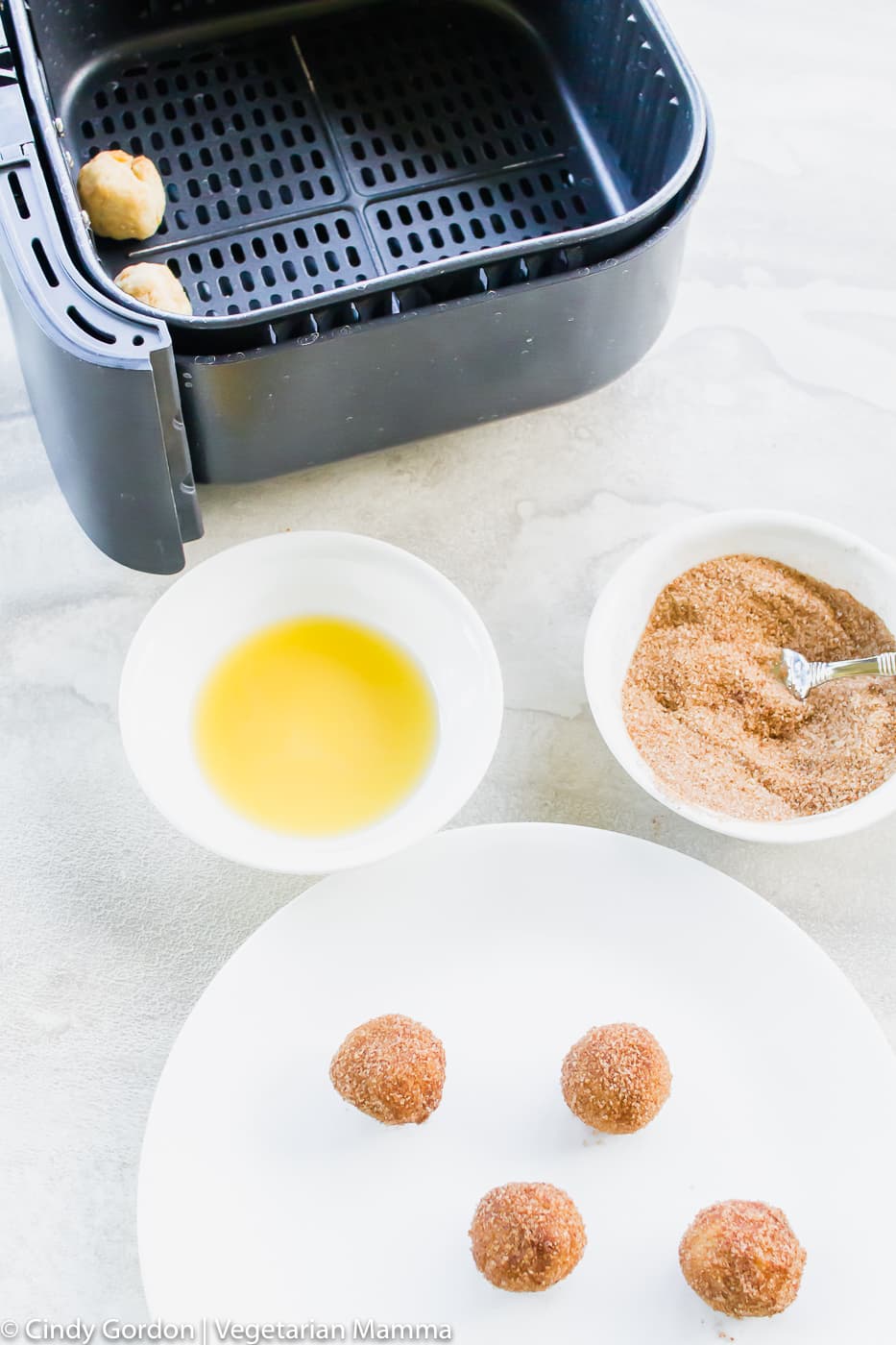 Air Fryer Donut Holes on white plate. Also pictured is a black air fryer basket and bowl of melted butter and sugar/cinnamon mixture.