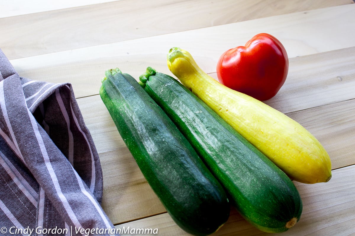 Two zucchinis, a yellow squash, and a tomato on a wooden cutting board. 