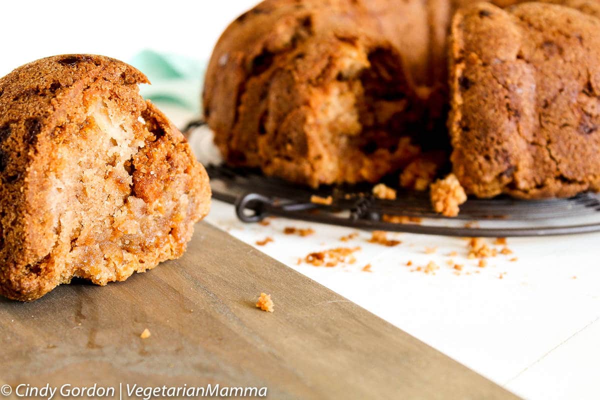 a slice of moist, tender apple bundt cake on a wooden board in front of the whole cake. 