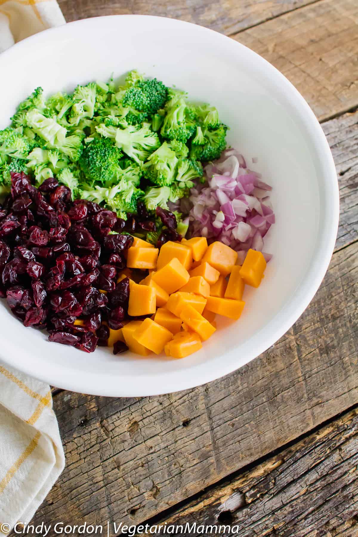 ingredients for broccoli salad in a bowl, including dried cranberries, yellow cheddar cheese, broccoli florets, and diced red onion. 