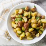 Top down view of roasted potatoes inside white bowl with white and yellow cloth beside bowl