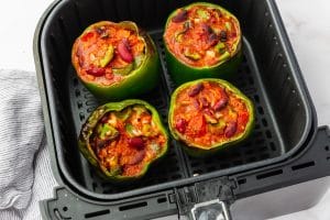 four green peppers stuffed with chunky red liquid in a back air fryer basket half cooked