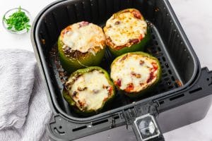 four green peppers stuffed with chunky red liquid in a back air fryer basket with melted white cheese on top