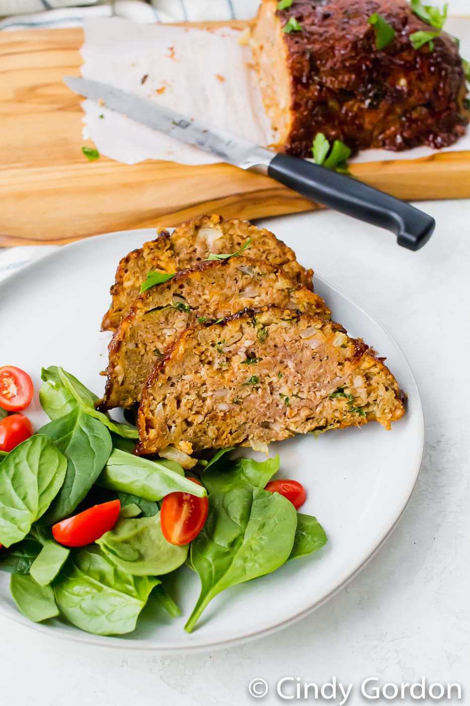 Air Fryer Meat Loaf sliced on a white plate with remaining loaf in the background. Served with green spinach and red tomatoes