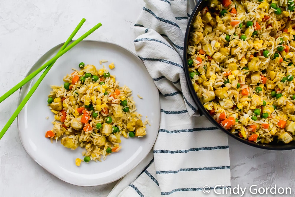 tofu fried rice with vegetables in a black skillet, aside a portion of the food on a round white plate with green chopsticks to the side 