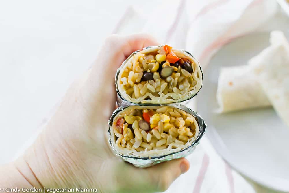 a hand holding two foil wrapped burritos filled with rice and veggies