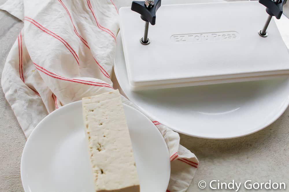 overhead picture of a block of tofu on a white plate and an EZ tofu press