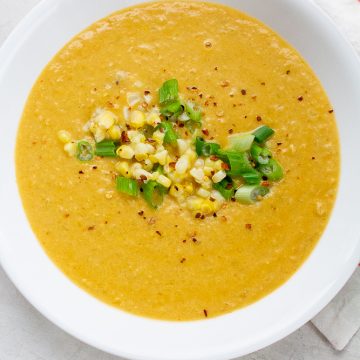 a bowl of yellow orange soup garnished with corn, scallions, and spices