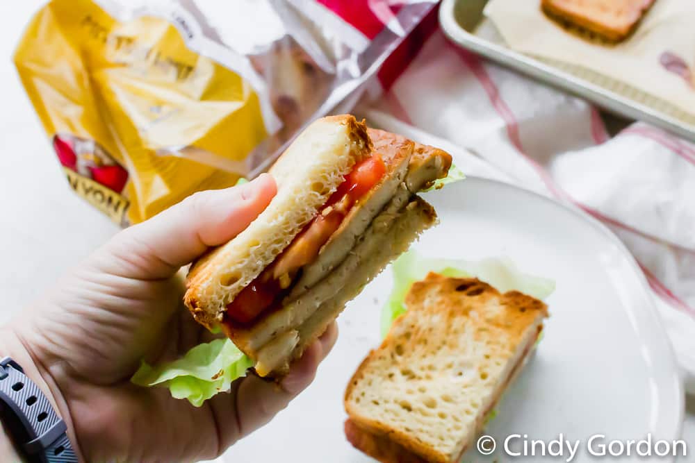 tofu bacon, lettuce, and tomato on toasted bread in a hand over top of a white place with a bag in the back ground