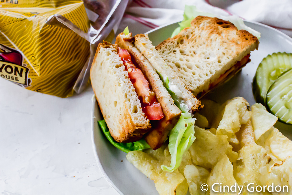 upclose picture of gluten free tofu bacon sandwich with chips and pickles on a white plate