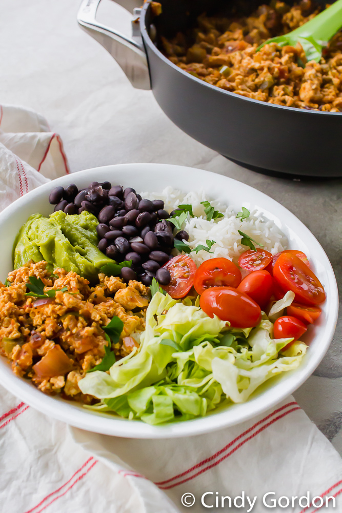 sofritas, lettuce, tomatoes, rice, black beans, and guacamole in a white bowl on a red and white towel.