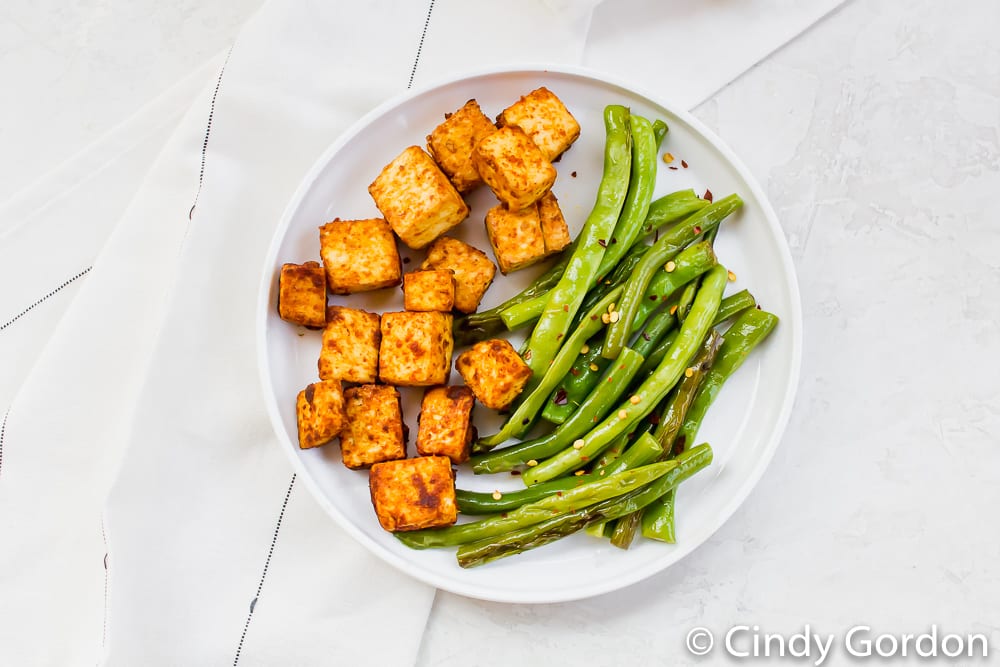 a round white plate on a marble countertop with roasted tofu and green beans