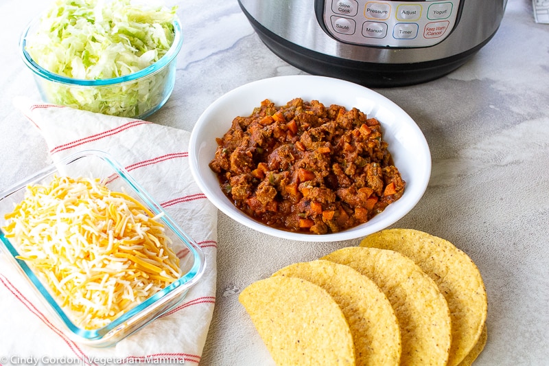 A white bowl of vegetarian taco meat is surrounded by cheese, lettuce, and taco shells