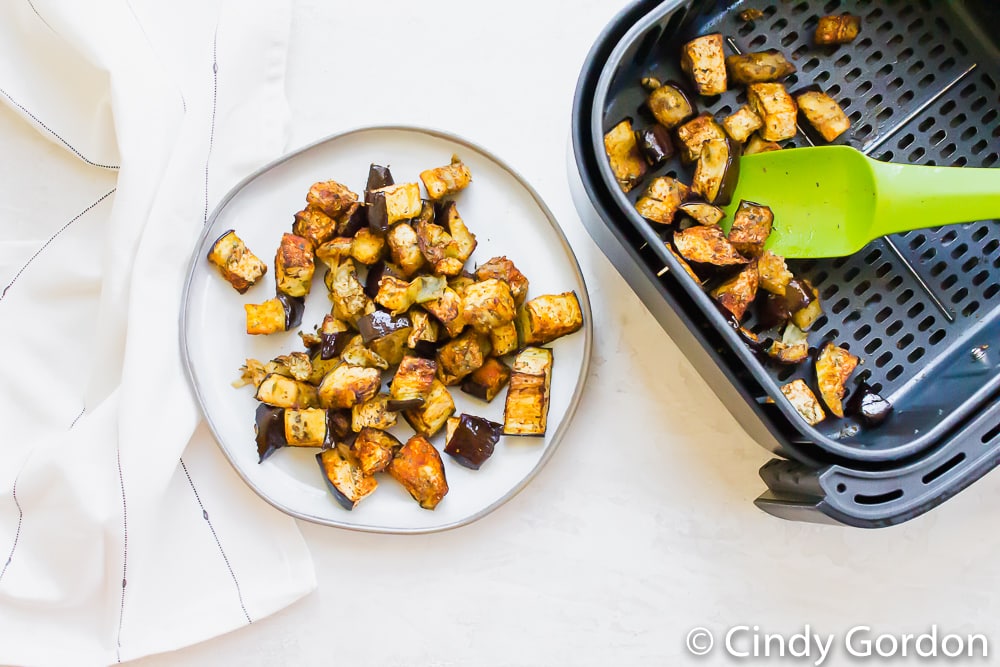 overhead view of air fryer eggplant on a white plate (that's on the left) on the right is a black air fryer basket with cooked eggplant cubes in side