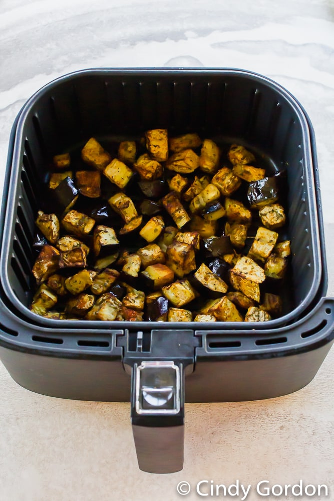 vertical photo of black air fryer basket with cooked eggplant cubes in it