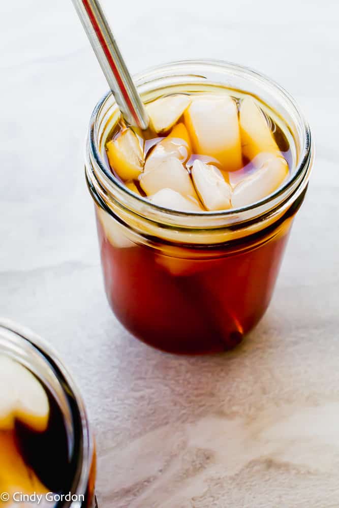 two pint mason jars filled with a brown liquid and ice cubes. One glass has a metal smoothie straw
