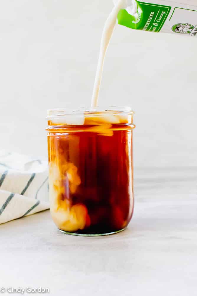vertical photo of brown coffee in a mason jar and white liquid being poured into the jar