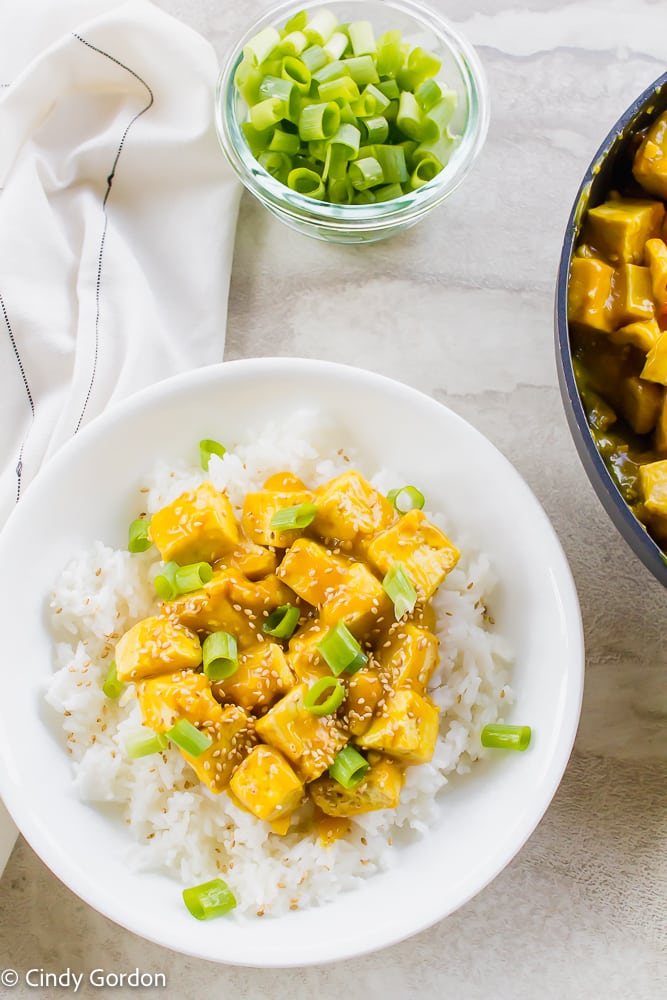 vegan orange chicken served over rice in a white bowl on a white back ground. Diced green onions in a round clear bowl at the top of the vertical picture