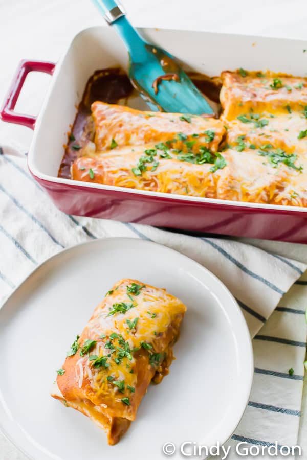 An enchilada on a round, white plate in front of a baking dish of enchiladas