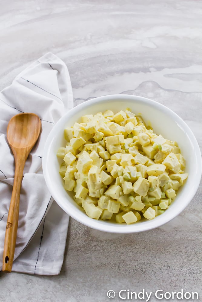 A white bowl of potato salad on a countertop with a wooden spoon and kitchen towel