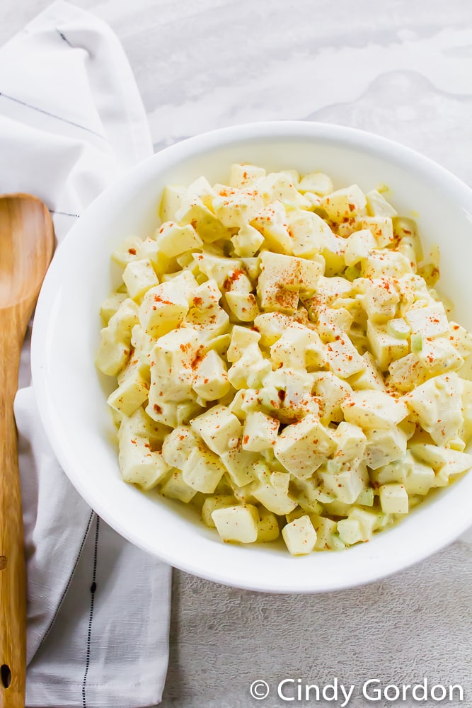 A white bowl of yellow potato salad garnished with paprika next to a wooden spoon