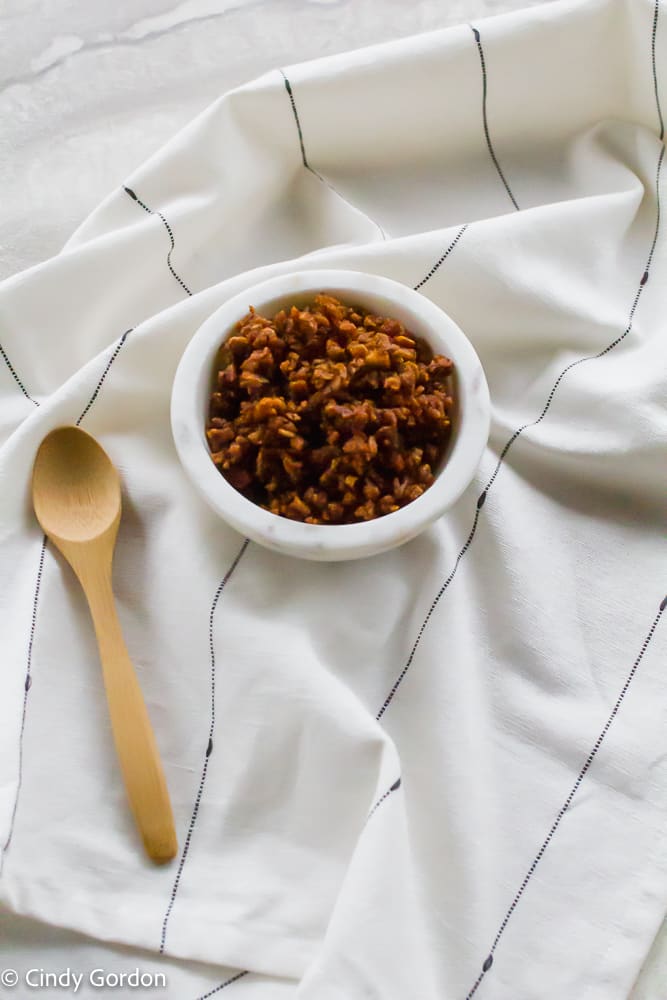 A small white bowl of bacon bits on a black and white kitchen towel with a wooden spoon