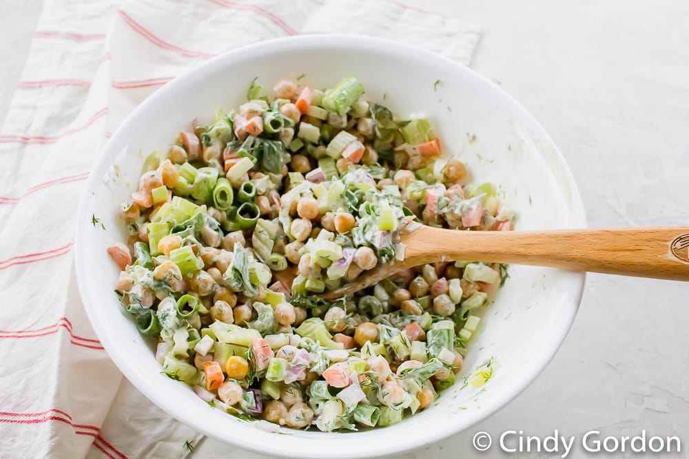 A white mixing bowl with chickpeas, carrots, celery, scallions, and a wooden spoon