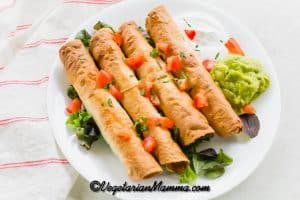 A white plate of taquitos with tomatoes, guacamole, sour cream, and cilantro