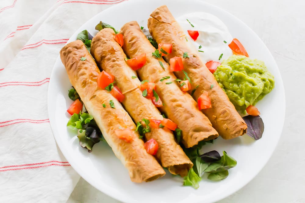 A white plate of taquitos with tomatoes, guacamole, sour cream, and cilantro