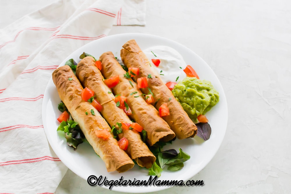 A white plate of taquitos with tomatoes, spring mix, guacamole, and sour cream
