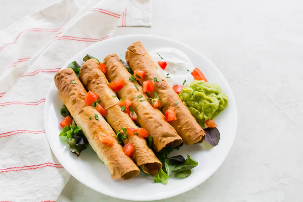 A white plate of taquitos with tomatoes, spring mix, guacamole, and sour cream