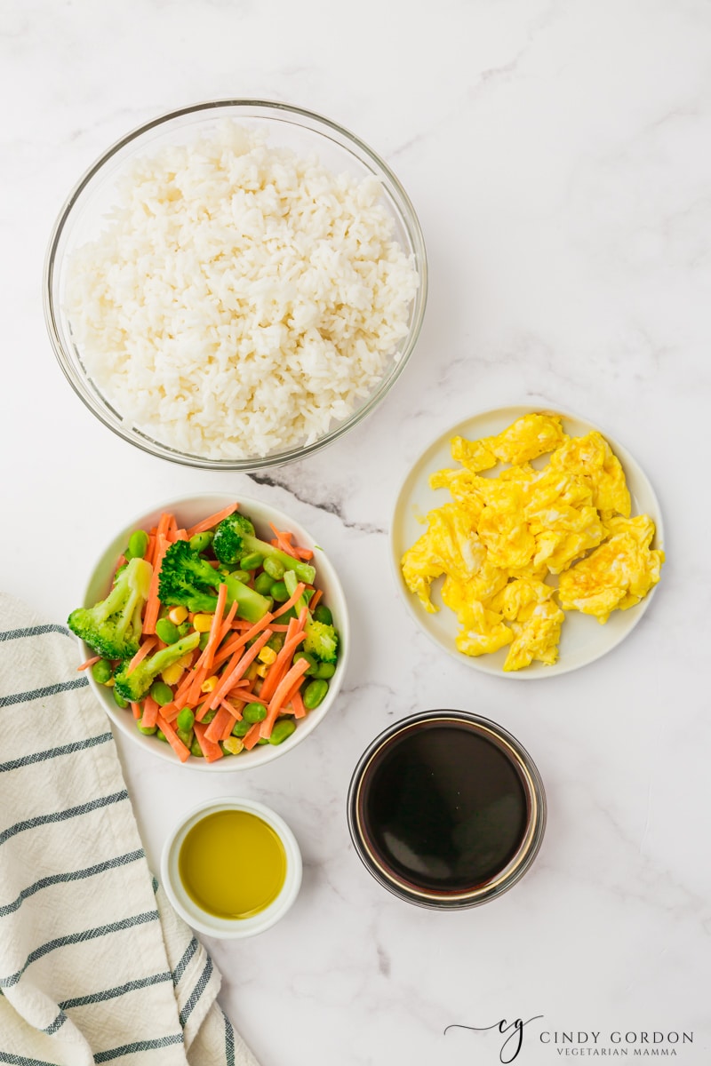 white background, bowl of white rice, mixed frozen veggies, scrambled eggs, brown liquid and yellow liquid.