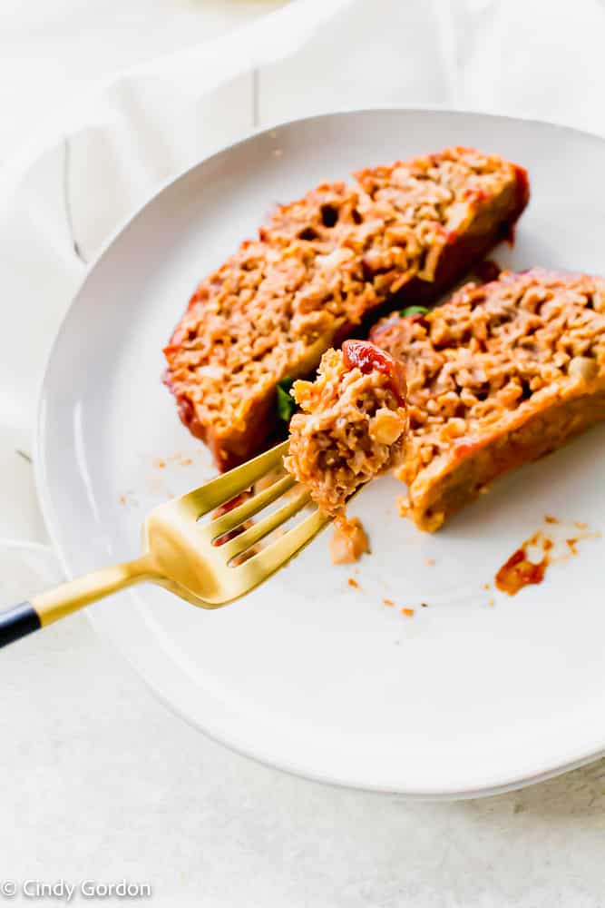 A bite of vegan meatloaf on a gold and black fork over a white plate of meatloaf