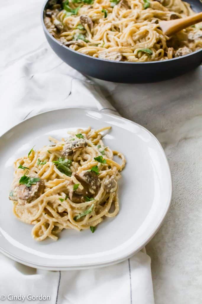 A white round plate of vegan carbonara with mushrooms next to a skillet of noodles