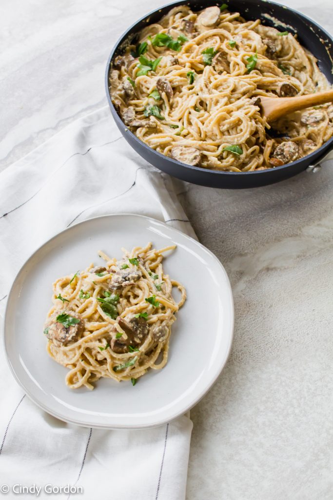 Overhead shot of vegan carbonara on a white round plate next to a sauté pan of noodles