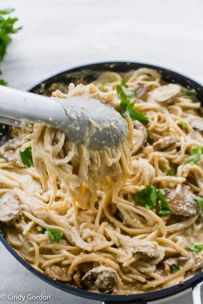 Metal tongs stirring spaghetti and mushrooms in vegan carbonara sauce to coat them evenly