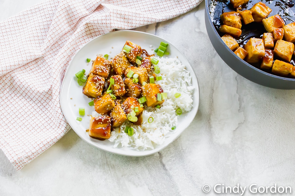 A plate of sesame tofu with white rice and green onions next to a pot of more sauced tofu