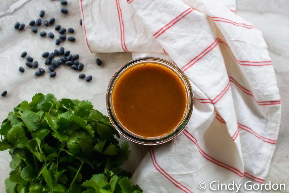 Overhead shot of a jar of homemade red enchilada sauce with a bunch of cilantro and a red striped kitchen towel