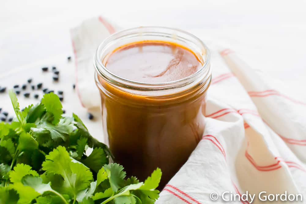 A jar of shiny enchilada sauce next to a red and white kitchen towel and a bunch of fresh cilantro