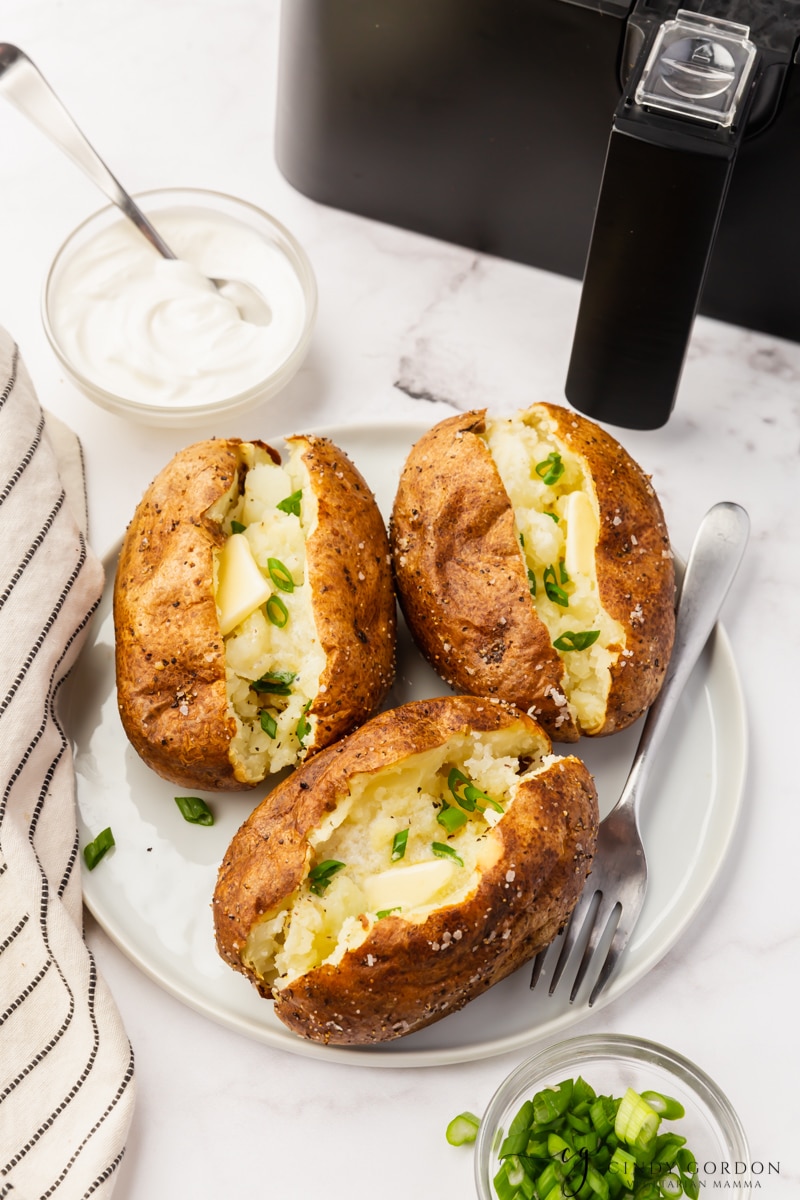 white back ground, black air fryer in top of pictures. Three baked potatoes, sliced open on a white plate with fork, Potatoes have sourcream and parsley