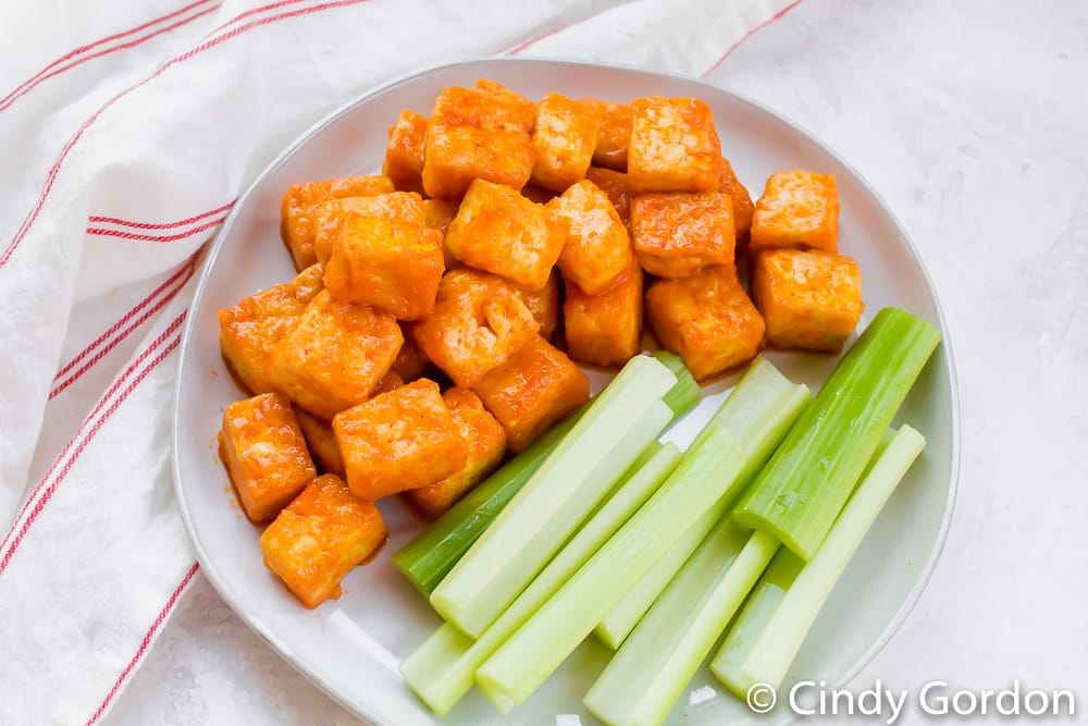 Bite-size pieces of tofu with buffalo sauce on a round white plate with celery sticks