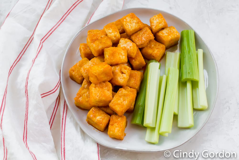 Cubed tofu covered in buffalo sauce alongside celery sticks on a white plate