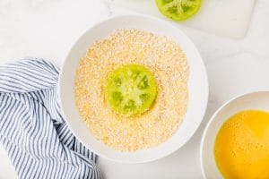 white bowl with corn meal mixture and a sliced green tomato with egg wash in the cornmeal
