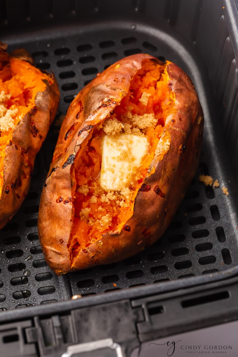 two air fryer baked sweet potatoes cut open with a square of butter and cinnamon on top. placed in a black air fryer basket