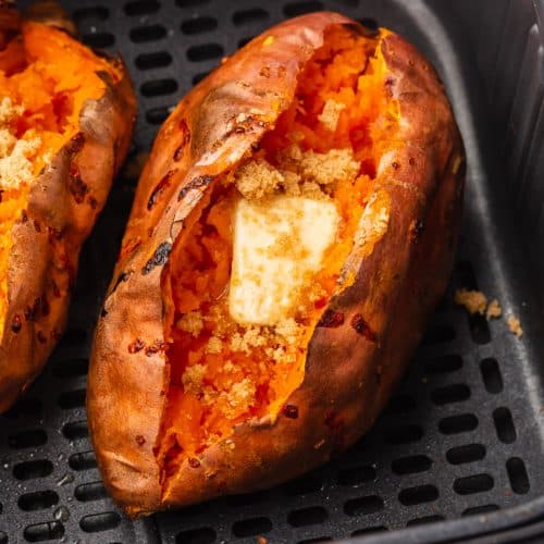 two air fryer baked sweet potatoes cut open with a square of butter and cinnamon on top. placed in a black air fryer basket