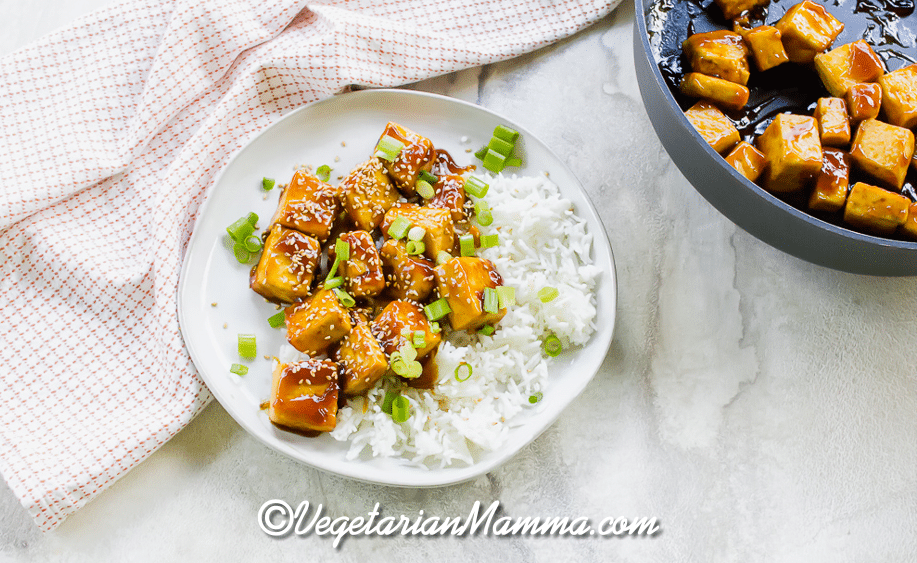 Overhead shot of a plate of sticky, crispy sesame tofu with white rice and sesame seeds with a pot of tofu