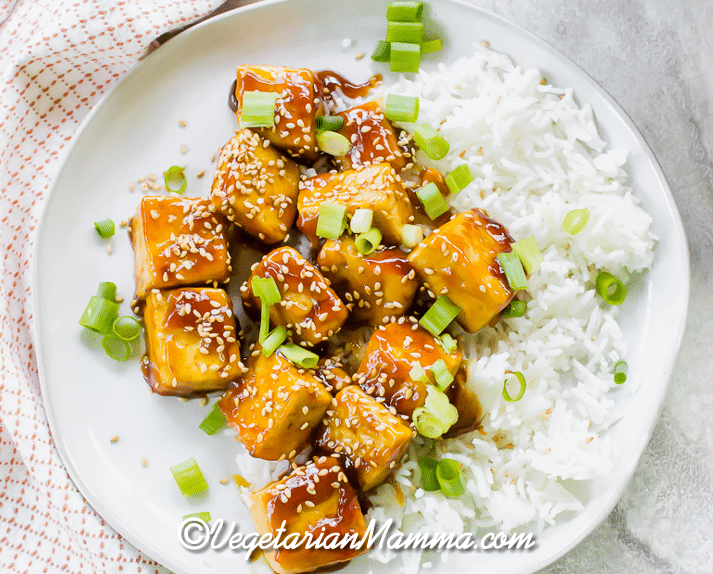 Overhead shot of sauced crispy tofu over white rice with sesame seeds and green onions on top.