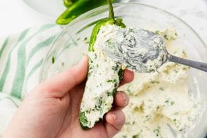 hand holding a sliced jalapeno pepper and a spoon stuffing it with white and green cream cheese