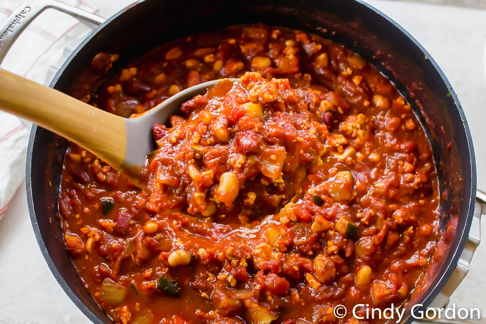 a ladle of vegetarian chili above the soup pot filled with tomato sauce, beans, tofu, and zucchini