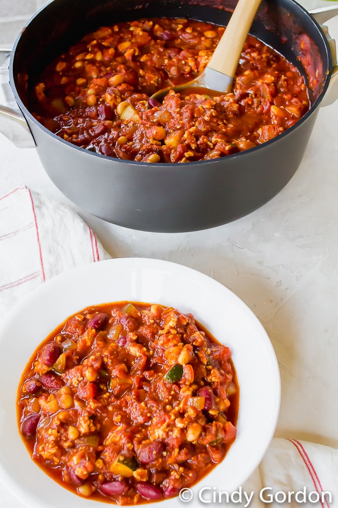 A round white bowl of tofu chili in front of a large pot filled with chili